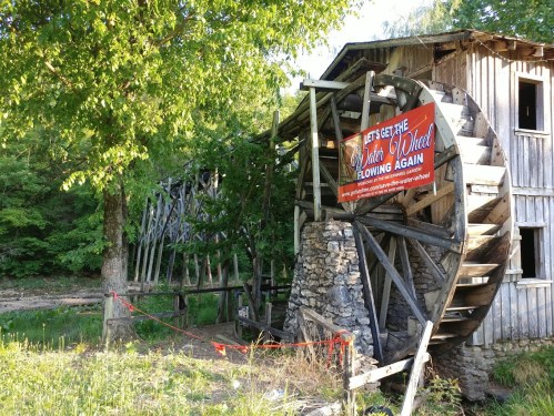 A wooden water wheel beside a rustic building, with a banner reading "Let's get the Water Wheel flowing again."