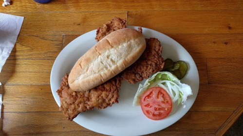 A plate with two fried chicken fillets, a sandwich bun, lettuce, tomato, and pickles on a wooden table.