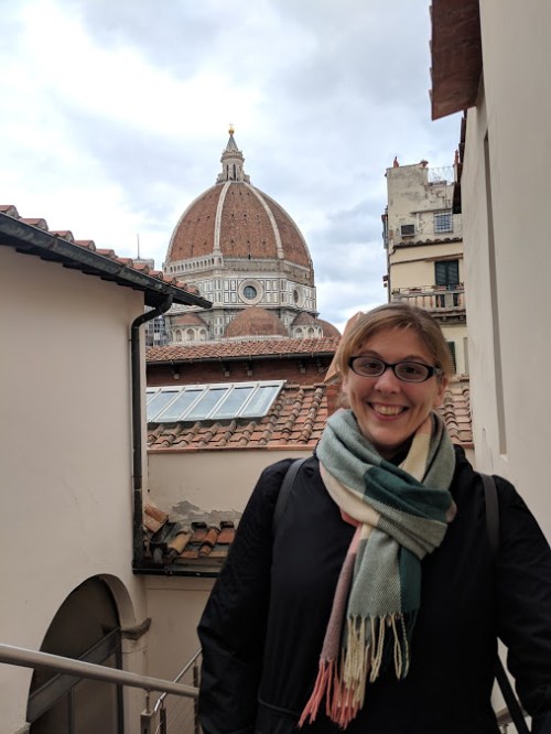 A smiling person stands in a narrow passageway with a view of a large dome in the background, surrounded by rooftops.
