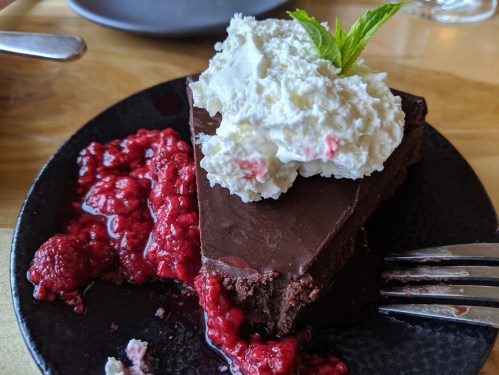 A slice of chocolate cake topped with whipped cream and a sprig of mint, served with raspberry sauce on a black plate.