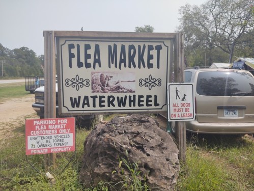 Sign for "Flea Market Waterwheel" with parking rules and a notice for dogs on a leash, near parked vehicles.