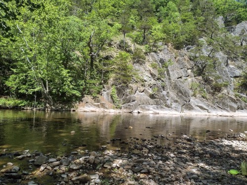 A serene riverbank scene with smooth rocks, clear water, and lush green trees lining a rocky hillside.