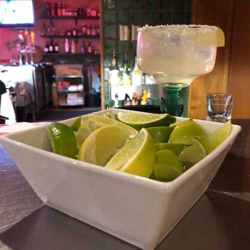 A bowl of lime wedges sits on a table next to a margarita glass in a vibrant bar setting.