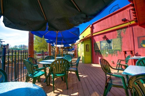 Outdoor seating area with green tables and chairs under blue umbrellas, colorful walls, and a wooden deck.