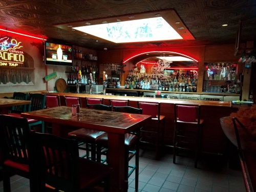A cozy bar interior with a wooden counter, empty tables, and a variety of bottles displayed on shelves. Neon lights glow.