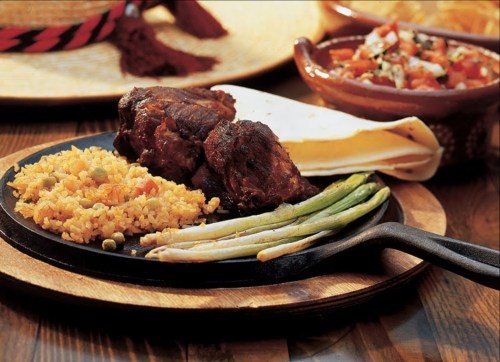 A sizzling plate with grilled meat, rice, green onions, and tortillas, accompanied by a bowl of salsa.