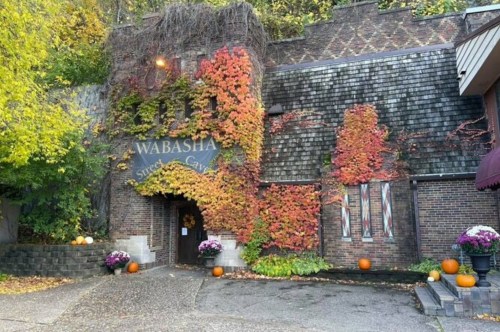 A charming brick building covered in colorful ivy, with pumpkins and flowers at the entrance, set in a scenic autumn landscape.
