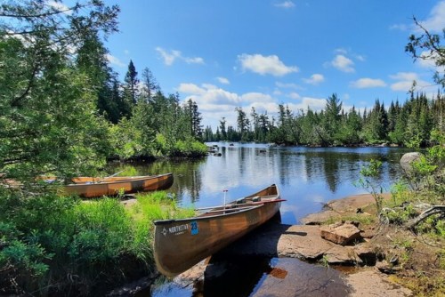 Two canoes are moored by a serene lake surrounded by lush greenery and trees under a bright blue sky.