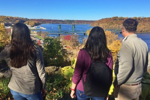 Three people stand overlooking a river and bridge, surrounded by autumn foliage and a clear blue sky.