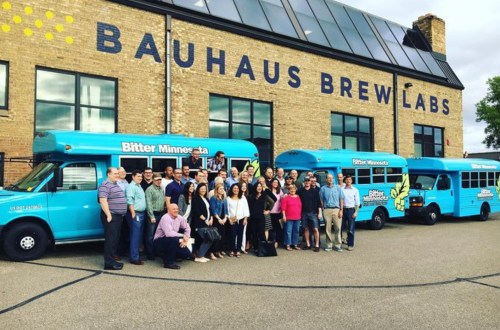 A large group of people poses in front of two blue buses at Bauhaus Brew Labs, a brewery with a brick building.