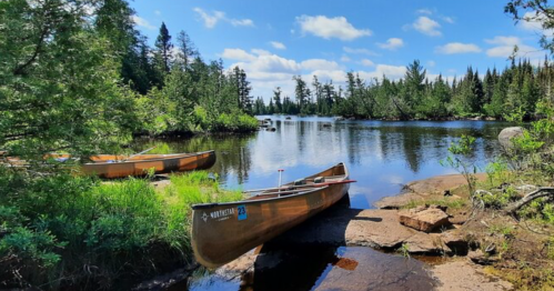 Two canoes on a rocky shore beside a calm lake, surrounded by lush greenery and a clear blue sky.