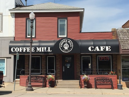A red cafe building with a sign reading "Coffee Mill Cafe" and an "Open" sign in the window. Flower pots are outside.