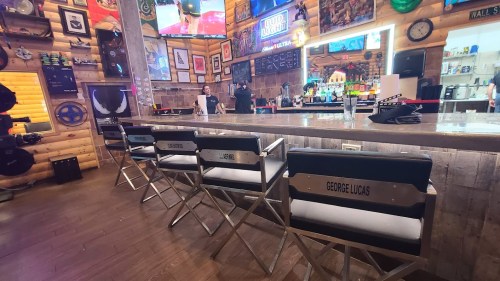 A bar with director's chairs labeled with names, including "George Lucas," in a rustic, decorated setting.