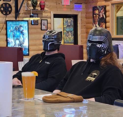 Two people wearing Kylo Ren masks sit at a bar, with a drink in front of one and a cozy atmosphere in a wooden setting.