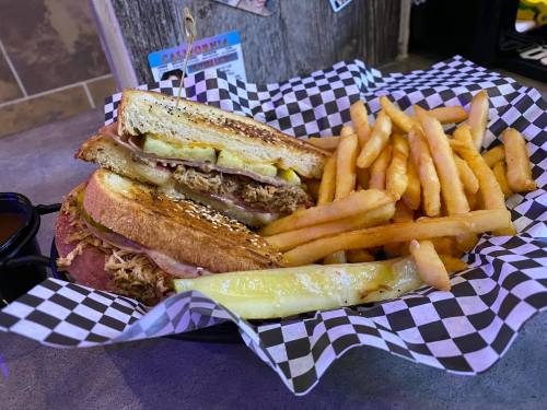 A sandwich with pulled pork and pickles, served with a side of crispy fries in a checkered basket.