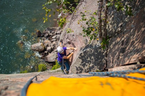 A climber ascends a rocky cliff near a river, with trees and water visible below.