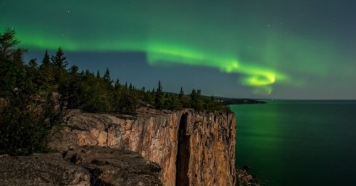 A stunning night scene featuring the Northern Lights over a rocky cliff and calm waters, surrounded by trees.