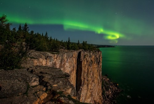 A stunning view of cliffs under a vibrant green aurora borealis illuminating the night sky over a calm sea.