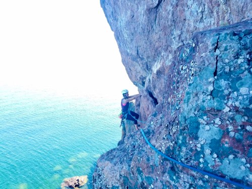 A climber ascends a rocky cliff by the sea, wearing a helmet and harness, with water visible below.