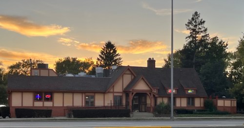 A cozy restaurant with a sloped roof, illuminated signs, and a sunset backdrop. Trees line the background.