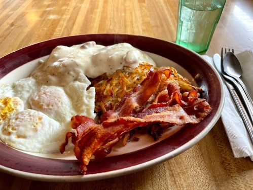 A plate of breakfast featuring eggs, bacon, hash browns, and biscuits with gravy, alongside a glass of water.