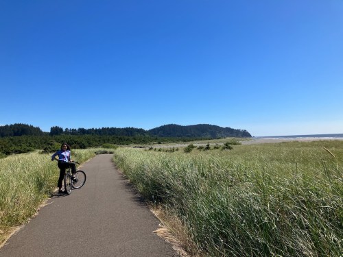 A person stands beside a bicycle on a path through tall grass, with a clear blue sky and coastline in the background.