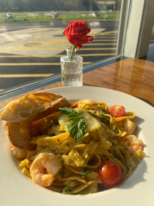 A plate of shrimp pasta garnished with herbs and cherry tomatoes, accompanied by a slice of bread and a rose in a vase.