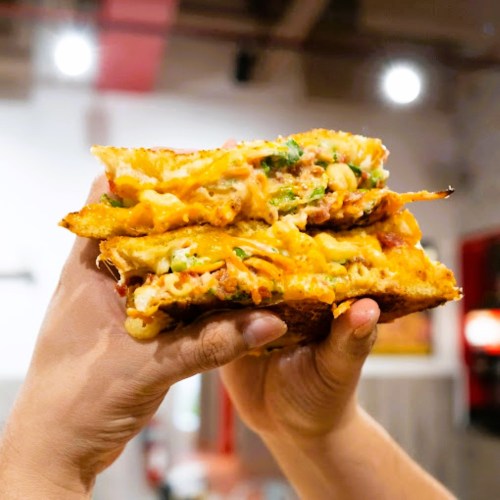 A person holding a stacked, cheesy sandwich filled with vegetables and meats, showcasing layers of melted cheese.