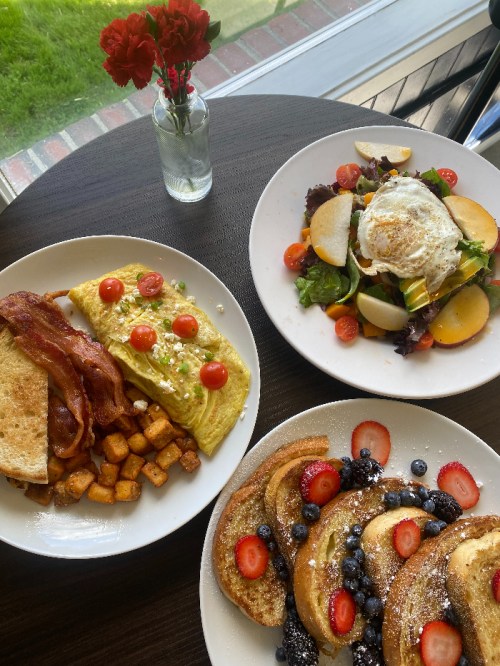 A table with three plates: a bacon omelet, a fresh salad with a fried egg, and French toast topped with berries.