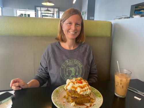 A woman sits at a table with a large dessert topped with whipped cream and a drink beside her, smiling at the camera.