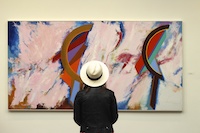 A person in a white hat stands before an abstract painting with colorful shapes and soft brush strokes.