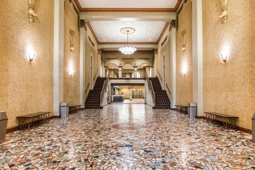 A spacious lobby with a mosaic floor, grand staircase, and elegant lighting, featuring textured walls and decorative details.