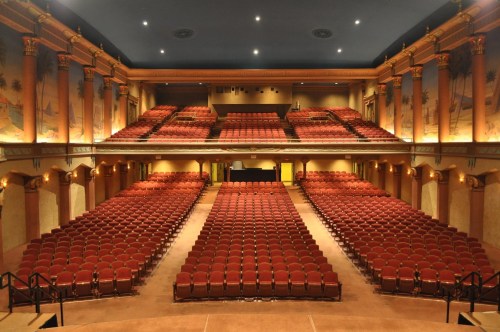 Interior of a theater with rows of red seats and decorative walls, viewed from the stage.