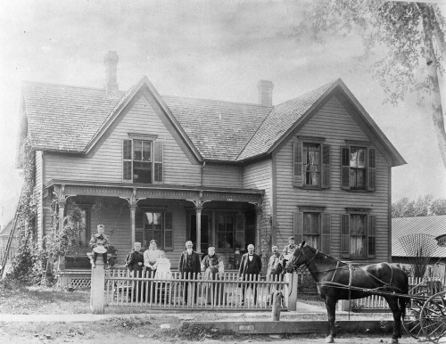 A historic black-and-white photo of a large house with a group of people and a horse-drawn carriage in front.