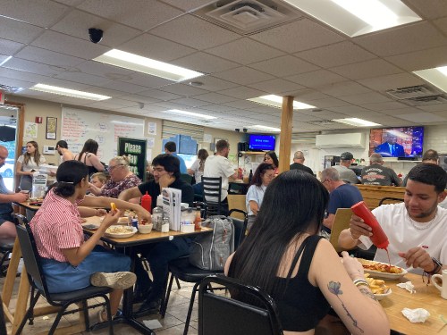 A busy diner scene with people eating at tables, chatting, and a TV playing in the background.