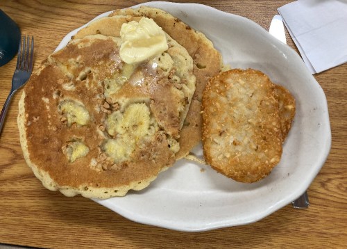 A plate with two banana pancakes topped with butter and a crispy hash brown on the side.