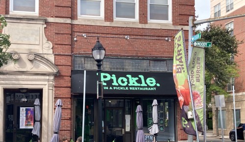 Exterior of a restaurant named "Pickle" with a green sign, located on a street corner with flags and outdoor seating.