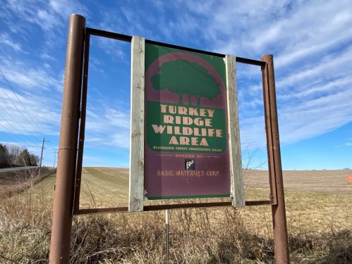 Sign for Turkey Ridge Wildlife Area, featuring green and maroon colors, with a blue sky and open field in the background.