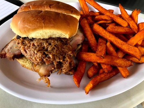 A pulled pork sandwich on a bun served with a side of crispy sweet potato fries on a white plate.