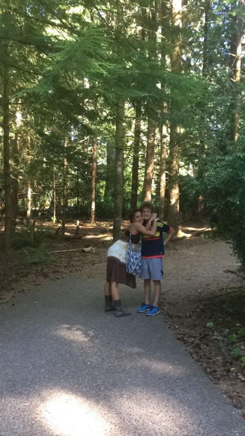Two children embrace on a forest path, surrounded by tall trees and greenery. Sunlight filters through the leaves.