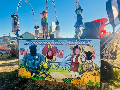 Two children pose behind a colorful cutout featuring cartoon firefighters and police, with pumpkins and hay bales in the background.