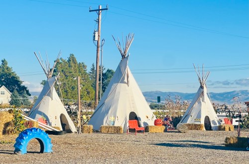 Three teepees with wooden poles, surrounded by hay bales and a blue tire, set against a clear sky and distant mountains.