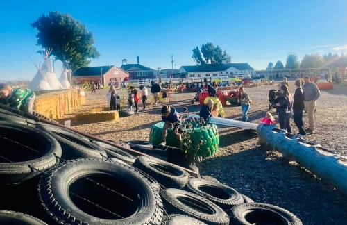 A vibrant outdoor scene with families enjoying a fall festival, featuring hay bales, tire stacks, and various activities.