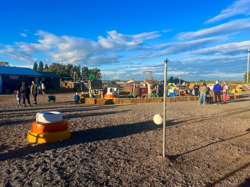 A lively outdoor play area with children and families, featuring colorful structures and hay bales under a blue sky.