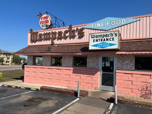 Exterior of Wampach's restaurant with a pink facade, sign reading "Fine Food" and "Entrance" for takeout. Clear blue sky.