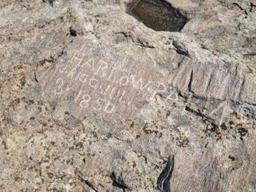 Carved inscription on a rocky surface, reading "Harlompa, 1850," with some moss and a small puddle nearby.