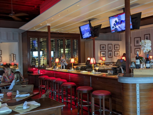 A stylish restaurant bar with red stools, wine racks, and TVs, featuring diners enjoying their meals in a cozy atmosphere.