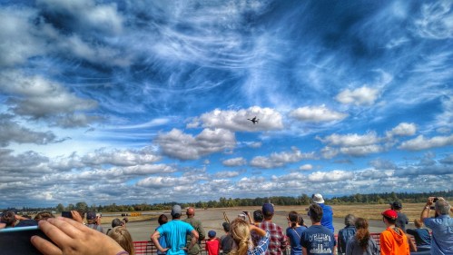 A crowd watches an aircraft perform in the sky, with dramatic clouds and a scenic landscape in the background.