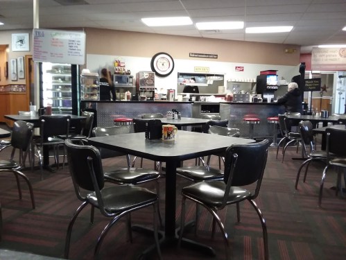 A diner interior with empty tables, a counter in the background, and a few patrons in a casual setting.