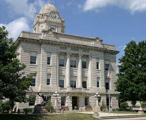 Historic stone courthouse with a clock tower, surrounded by trees and a grassy area under a blue sky.